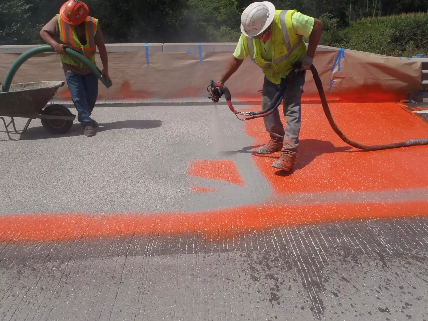 An image of two construction workers using a sprayer to apply an orange coating to a bridge deck.